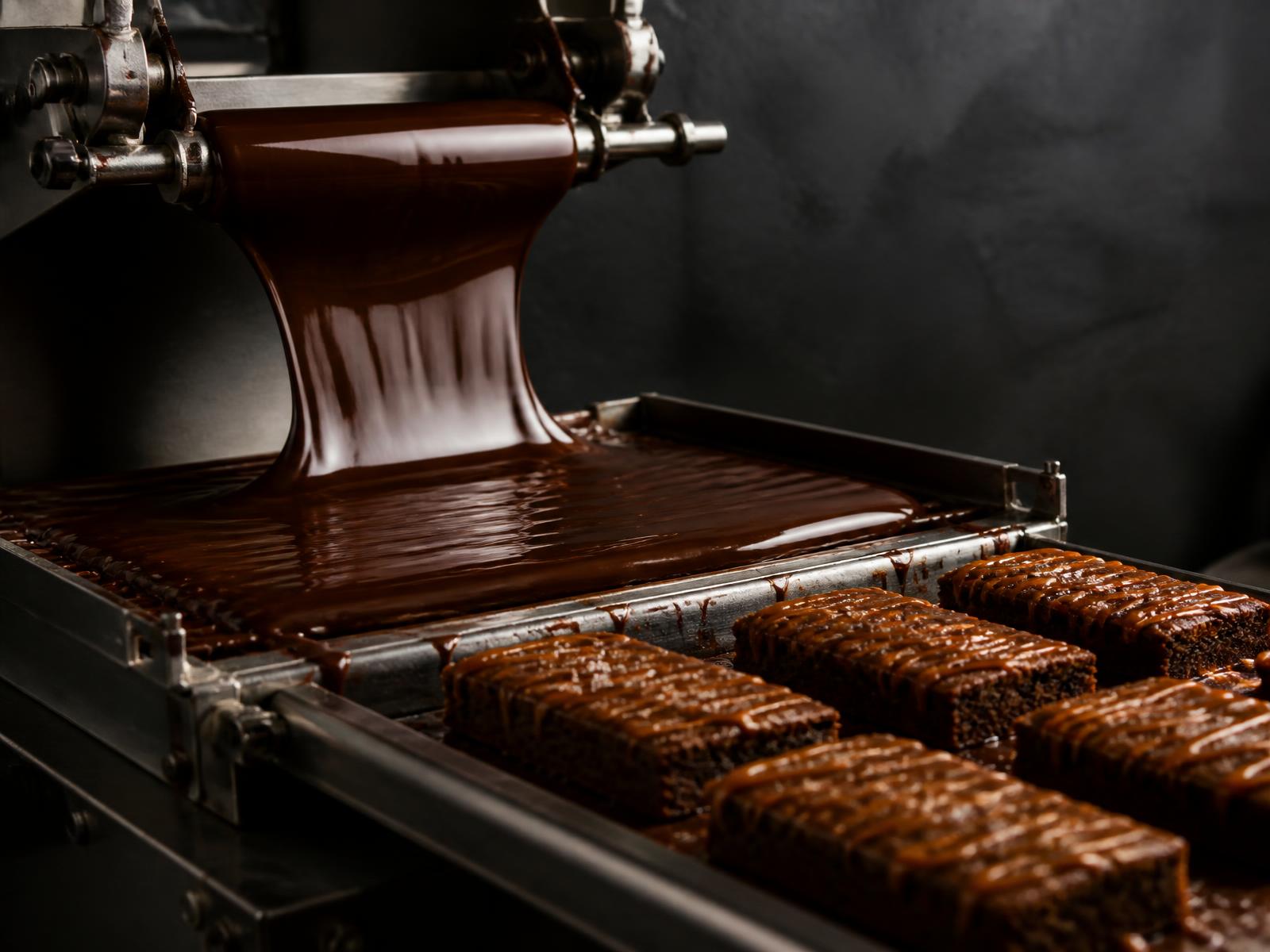 Melted chocolate pouring onto a production line beside trays of caramel-drizzled brownies